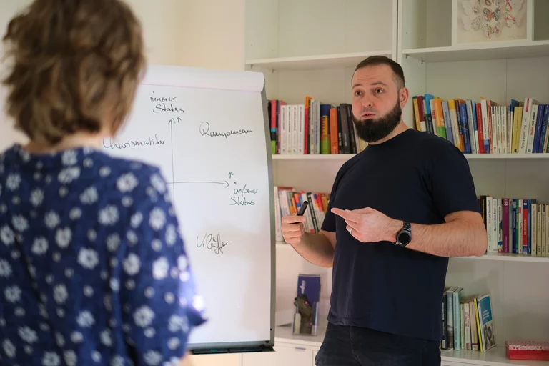 Dr. Sebastian Hümbert-Schnurr steht vor einer Flipchart und erläutert eine Skizze, eine Klientin steht ihm zugewandt im Vordergrund.
